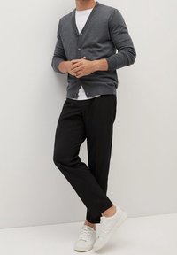 Mann trägt graue Strickjacke, weißes T-Shirt, schwarze Hose und weiße Turnschuhe, steht mit locker verschränkten Händen an einer weißen Wand.