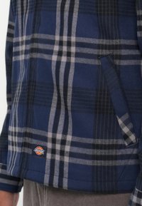 Camisa de flanela xadrez azul marinho com riscas cinzas e pretas, um bolso lateral e uma etiqueta de logo Dickies junto à bainha. Textura macia.