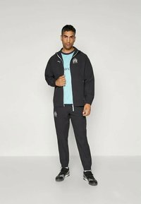 Mann står iført svart Puma treningsdrakt med logoene til Marseille fotballklubb, lys blå skjorte under, og svarte joggesko foran en ensfarget bakgrunn.