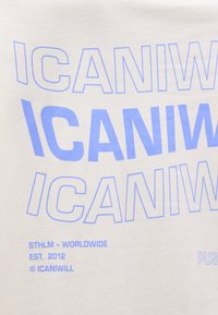 Επαναλαμβανόμενο μπλε κείμενο "ICANIWILL" με έντονα γράμματα και περίγραμμα σε εκρού ύφασμα, με μικρότερο κείμενο "STHLM - WORLDWIDE EST. 2012 © ICANIWILL" από κάτω.