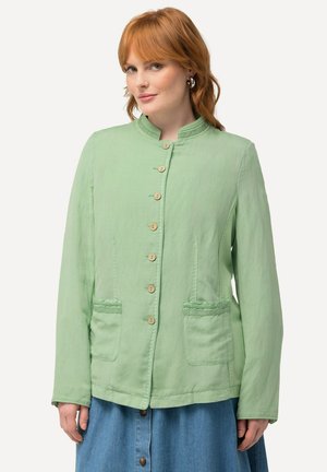 Giacca verde chiaro con colletto rialzato, bottoni in legno, due tasche frontali e tessuto testurizzato, abbinata a una gonna di jeans.