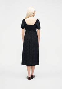 Γυναίκα με μακ ριά ξανθά μαλλιά φορώντας μαύρο midi φόρεμα με λευκές πουά και φουσκωτά μανίκια, στέκεται με την πλάτη σε απλό φόντο.