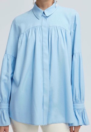 Jasnoniebieska bluzka z długim rękawem z marszczonym karczkiem i rękawami, ukrytymi guzikami z przodu oraz spiczastym kołnierzem, noszona z białymi spodniami.