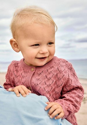 Małe dziecko o jasnych włosach, ubrane w różowy, dziergany sweterek, uśmiechające się i opierające się o osobę w niebieskiej koszuli na plaży.