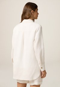 Camicia di lino bianca con maniche lunghe, polsini a bottone e piega sul retro. Tessuto morbido e vestibilità rilassata, abbinata a pantaloni corti leggeri.