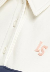Gros plan sur une chemise à col blanc avec des boutons et les lettres "LS" brodées en rose sur le tissu au-dessus d'une bordure bleu marine.
