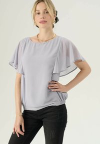 Una donna con capelli biondi raccolti indossa una blusa leggera con maniche a ruota di colore viola chiaro, pantaloni neri, orecchini a cerchio dorati e collane a strati, in piedi con una mano sui fianchi.