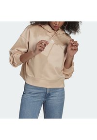 Ljust beige huvtröja med dragsko, i bomullsmaterial, och en liten vit logga på bröstet. Bärs tillsammans med blå denimjeans.