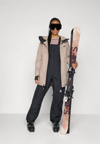 Snowboarder i en beige jakke og svarte overalls, holder en ski med bindinger. Store svarte briller, teksturert genser under, solide støvler.