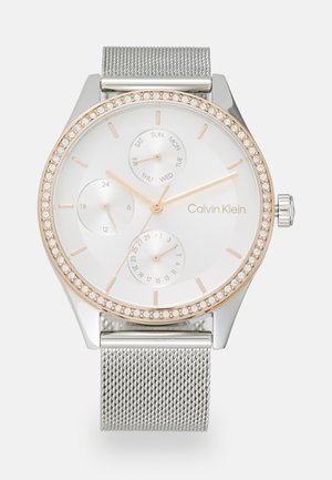 Reloj con correa de malla plateada y esfera blanca, bisel en tono dorado adornado con cristales, tres subesferas y un diseño minimalista.