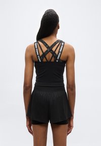 Γυναίκα που φοράει μαύρο αθλητικό σορτς και μαύρο αθλητικό τοπ με σταυρωτούς ιμάντες με την επωνυμία PUMA, στέκεται γυρισμένη προς τα πίσω σε λευκό φόντο.