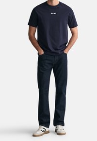 T-shirt a girocollo navy con logo bianco "GANT", abbinato a jeans blu scuro e sneakers chiare con dettagli verdi.