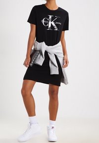 Svart T-shirt med silverfärgad Calvin Klein-logga, kombinerad med en grå sweatshirt knuten i midjan, svart kjol och vita sneakers.