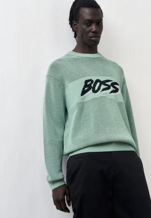 Uomo che indossa un maglione a rete verde chiaro con la scritta "BOSS" in nero e bianco, abbinato a pantaloni neri, in piedi su sfondo bianco.