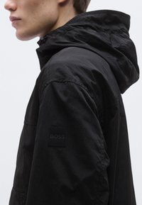 Must kerge jope, millel on kapuuts, matt tekstuur ja vasakul varrukal ruudukujuline logo märk. Disain on voolujooneline ja kaasaegne.