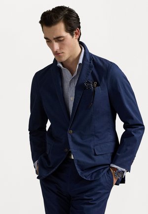 Marineblaues Baumwollblazer mit Reverskragen, Brusttasche und goldenen Knöpfen, kombiniert mit einem gestreiften blauen Hemd und einem gepunkteten Taschentuch.