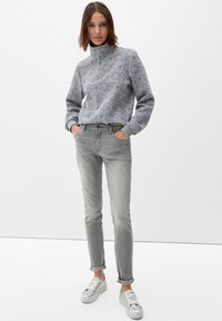 Sudadera gris con cremallera y un sutil patrón de logo, combinada con jeans slim de un gris claro y zapatillas plateadas, sobre un fondo blanco liso.