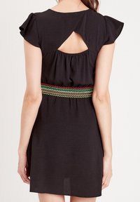 Schwarzes Kleid mit kurzen, leicht ausgestellten Ärmeln, einem Rückenausschnitt und einem elastischen Bund mit buntem Zickzackmuster in Rot, Grün und Gelb.