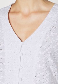 Nahaufnahme einer weißen Bluse aus Spitzenstoff mit kleinen Knöpfen auf der Vorderseite und einem V-Ausschnitt, der den Bereich des Schlüsselbeins zeigt.