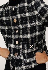 Sorte og hvide ternede jakke med tekstureret stof. Har guld- og sorte dekorative knapper samt et kraveløst design.