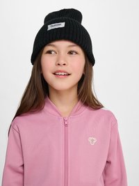 Dziecko noszące czarną dzianinową czapkę z metką "hummel sportswear" i różową zapinaną na zamek kurtkę z małym logo motyla, patrzące lekko na bok.
