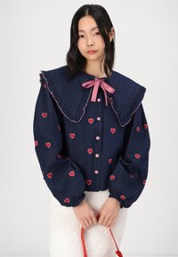 Τζιν μπλούζα με μεγάλο ριχτό γιακά, ροζ φιόγκο και κεντημένη κόκκινη καρδιά. Διαθέτει κουμπιά μπροστά και φουσκωτά μανίκια.