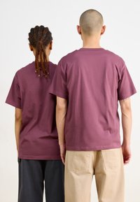 Des t-shirts bordeaux portés par deux personnes, avec des manches courtes et une coupe décontractée, associés respectivement à des pantalons beiges et gris foncé.