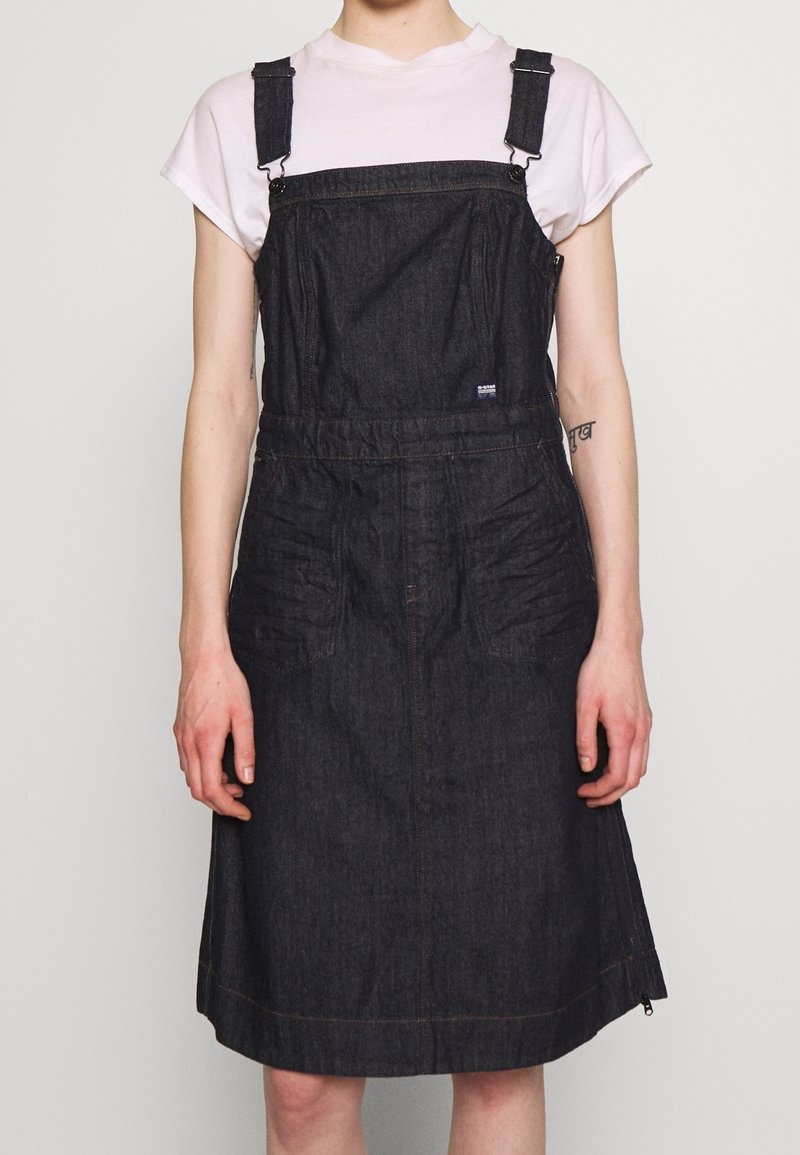 Robe salopette en denim foncé avec bretelles réglables, poches avant et ourlet coupé au niveau du genou. Présente des accents de surpiqûres discrètes.