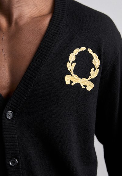 Svart cardigan i stickat material med en guld broderad lagerkrans och banddetalj på vänster bröst. Svarta knappar är synliga.