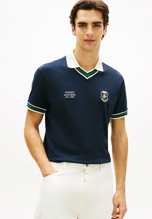 Joven con polo azul marino con cuello blanco y ribetes a rayas, pantalones blancos, de pie con una mano en el bolsillo.