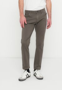 Vyras modelis dėvi rudas medvilnines chino kelnes, siauro kirpimo, su penkiomis kišenėmis, metaliniais vinutėmis ir subtilia tekstūra. Derinamos su baltais sportbačiais.