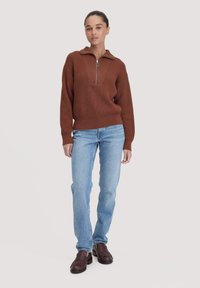 Rostfarbener, gerippter Pullover mit einem Viertelzip-Kragen, kombiniert mit hellblauen, geraden Jeans und braunen Slippern.
