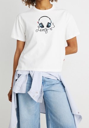 Person iført en hvid T-shirt med et høretelefon-grafik og lyseblå jeans, med en stribet skjorte bundet om livet.