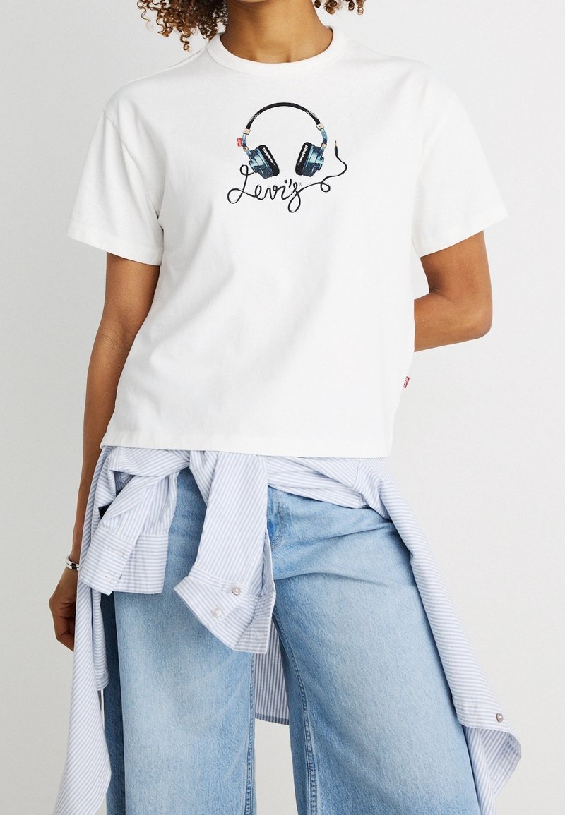 Personne portant un T-shirt blanc avec un motif de casque audio et un jean bleu clair, avec une chemise rayée nouée autour de la taille.