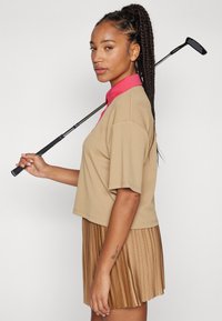 Μπεζ πουκάμισο γκολφ με ροζ γιακά, κοντά μανίκια και υφή, συνδυασμένο με έναν πλισέ καφέ φούστα. Κλαμπ γκολφ κρατημένο πάνω από τον ώμο.