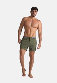 Shorts de bain verts avec une ceinture rayée, dotés d'une fermeture à bouton et de poches latérales, portés par un modèle masculin debout pieds nus.
