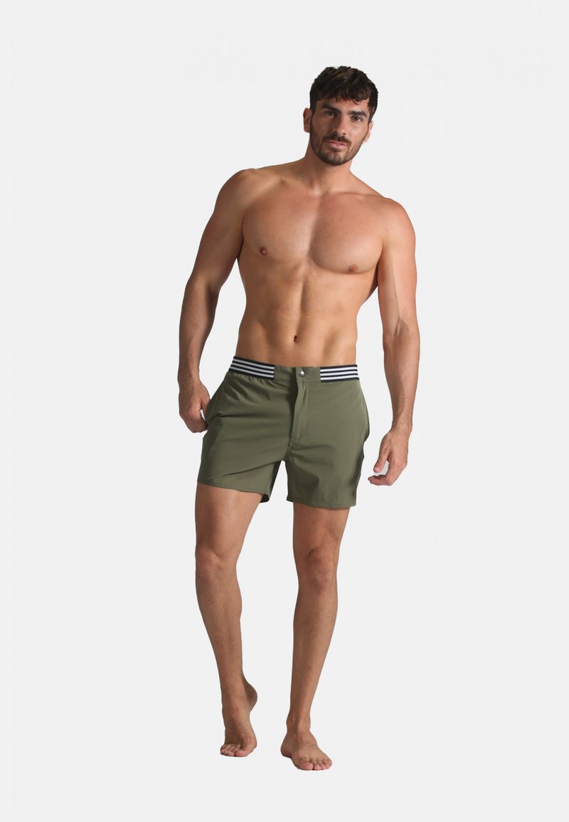 Shorts de bain verts avec une ceinture rayée, dotés d'une fermeture à bouton et de poches latérales, portés par un modèle masculin debout pieds nus.