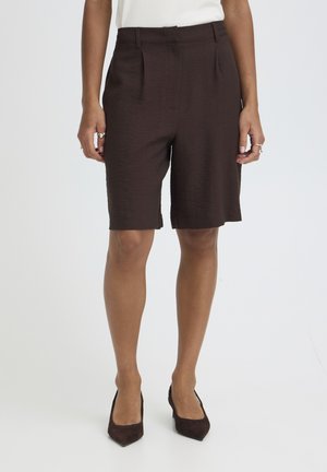 Kobieta ubrana w ciemnobrązowe, dopasowane bermudy do kolan, biały top i ciemnobrązowe szpilki z zaostrzonym czubkiem, stojąca na tle jednolitego tła.