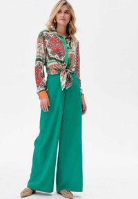 Kleurvolle blouse met een patroon en een geknoopt voorpand, gecombineerd met hoog-waisted, wijduitlopende groene broeken. Beige platte schoenen maken de outfit compleet.