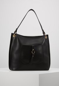 Czarna skórzana torebka na ramię z przednią kieszenią, ozdobiona paskiem i detalem w postaci złotego pierścienia, prezentowana na białym piedestale.