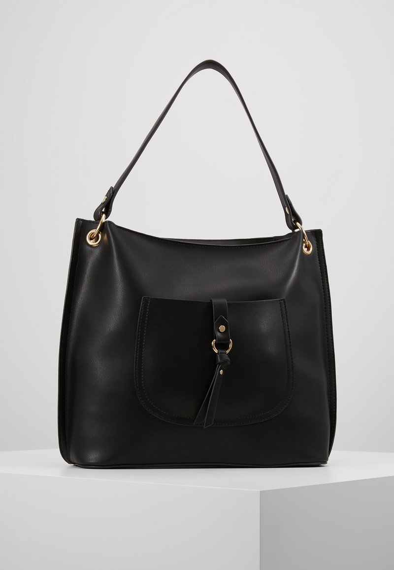 Czarna skórzana torebka na ramię z przednią kieszenią, ozdobiona paskiem i detalem w postaci złotego pierścienia, prezentowana na białym piedestale.