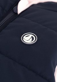 Tamsiai mėlynas pūkinis švarkas su šviesiai rožiniu apykakle. Yra apskritas logotipo ženkliukas su burlaivio piešiniu ir užtrauktuko uždarymas.