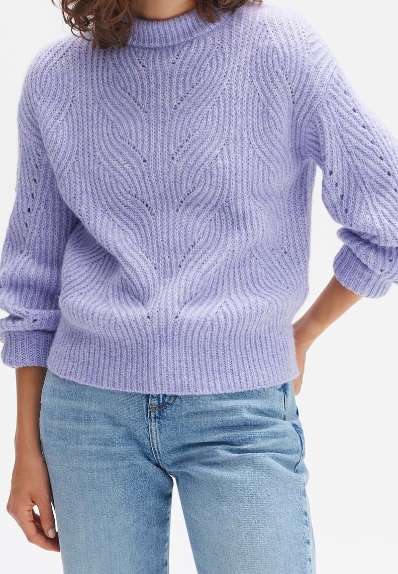 Kvinna iförd en lavendelfärgad kabelstickad tröja med uppvikta ärmar, tillsammans med ljusblå denimjeans, handen vilande på höften.