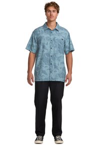 Young man standing, wearing a blue short-sleeve button-up shirt with a textured pattern, black pants, and black sneakers with white soles.