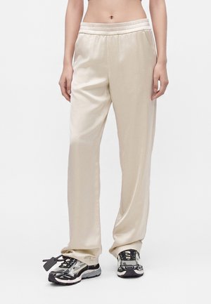 Person iført løstsiddende beige træningsbukser kombineret med sort-hvide atletiksneakers, stående på en ensfarvet lys baggrund.