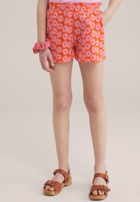 Rde kratke hlače z belim in rožnatim vzorcem marjetic, elastičnim pasom, v kombinaciji z rjavimi usnjenimi sandali z debelim petkom in zaponkami.