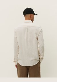 Lichtbeige langwerpige blouse met een textuurpatroon, knoopmanchetten en een afgeronde zoom. Heeft een rugpas en plooidetail.