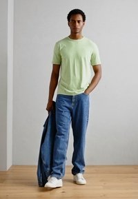 Tricou din bumbac verde deschis, blugi din denim albastru, relaxați, și adidași albi. Ținând o jachetă din denim albastru, stând pe un podea ușoară din lemn.