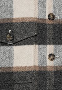 Geruit wollen overhemd met grijze, beige en bruine kleuren, twee knopen, een borstzak en een textuurstof met zichtbare stiksels.