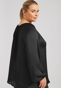 Svart långärmad blus gjord av strukturerat tyg med lös passform, rundad halsringning och samlad detalj vid axlarna.